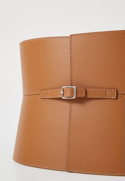 Ceinture en cuir tan avec une finition mate, présentant une petite boucle en argent et des accents piqués. Le design a une forme large et structurée.