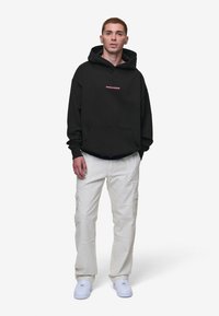 Svart hettegenser med lomme foran og "PEGAADOR" i rosa tekst. Kombinert med off-white cargo-bukser med sidelommer og hvite joggesko.