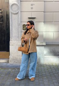 Jasnobrązowa skórzana kurtka z kołnierzem, połączona z jasnoniebieskimi dżinsami o szerokich nogawkach i beżowymi butami typu slip-on. Nosi małą brązową torebkę.