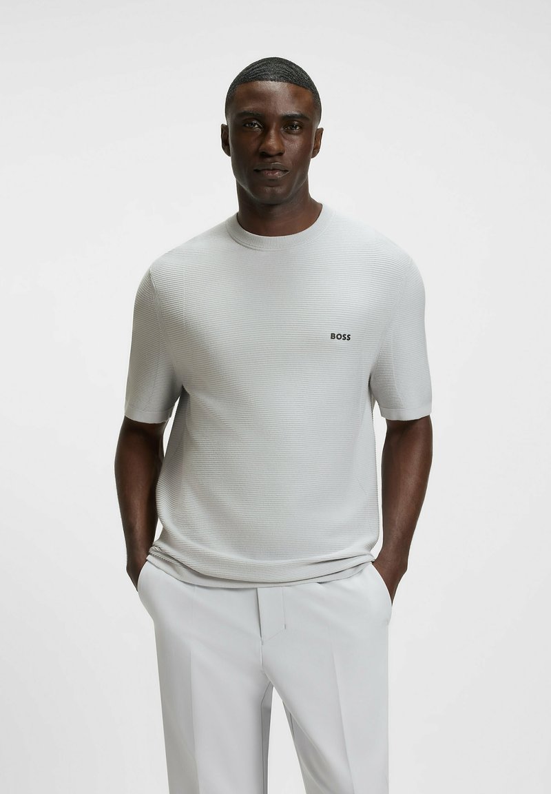 Light grey short-sleeve knitted sweater with a round neckline and small black "BOSS" logo on the chest, worn with light-colored trousers.