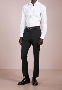 Bílá košile s dlouhými rukávy spárovaná s černými kalhotami na míru a černými formálními botami. Má elegantní, minimalistický design s akcentem pásku.