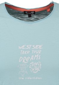 Camisa de manga corta azul claro con texto impreso en blanco. El texto dice "LADO OESTE, TOMA TUS SUEÑOS", con elementos gráficos y logotipo.