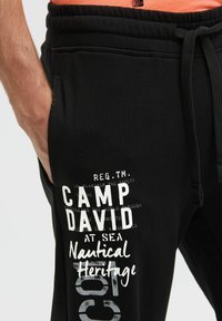 Schwarze Jogginghose mit einem elastischen Bund und Kordelzug. Mit weißem Grafiktext "CAMP DAVID AT SEA Nautical Heritage". Weiche Textur.