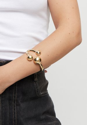 Brazalete de oro con un diseño fluido, que presenta dos acentos esféricos lisos colocados asimétricamente a lo largo de la banda.