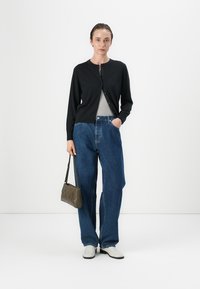 Cardigan nero su una maglia grigia, abbinato a jeans blu larghi. Il modello tiene una borsa marrone e indossa scarpe bianche. Design semplice, stile casual.