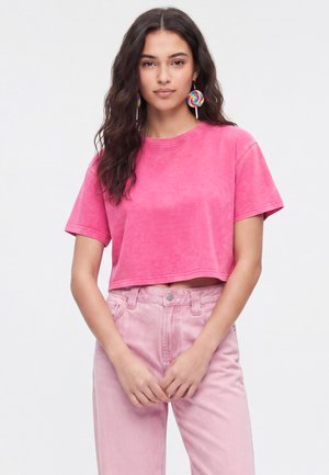 Młoda kobieta o długich falujących włosach nosi różowy krótki t-shirt, jasnoróżowe spodnie z wysokim stanem i kolorowe kolczyki w kształcie lizaków na białym tle.