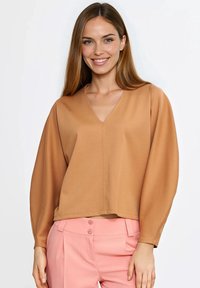 Brązowa bluzka z długim rękawem w serek, wykonana z gładkiej tkaniny; charakteryzuje się luźnym krojem i minimalistycznym wykończeniem szwów. W zestawie z różowymi spodniami.