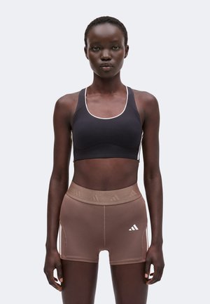 Mujer atlética de pie de frente, lleva un sujetador deportivo negro y pantalones cortos deportivos marrones de cintura alta con logo sobre fondo blanco.