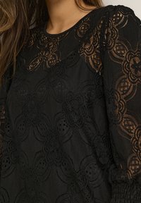Top en dentelle noire avec des motifs floraux, des accents transparents et un tissu texturé. Manches longues avec des poignets froncés et un col rond.