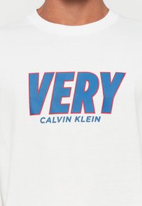 Balta marškinėliai su dideliu mėlynu užrašu "VERY" raudona apvadu, o po juo ant krūtinės mažesniu mėlynu užrašu "Calvin Klein".