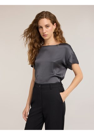 Frau mit langen welligen Haaren, die ein graues Satin-Kurzarmshirt und schwarze hoch taillierte Hose trägt, stehend mit einer Hand in der Tasche.