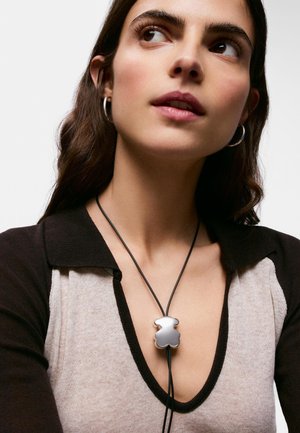 Joven mujer con un top beige y negro con escote en V pronunciado, pendientes de aro y un colgante plateado con forma de oso en un collar de cordón negro.