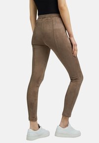 Vrouw in strakke bruine suedine leggings en witte sneakers, staande tegen een witte achtergrond, gezien vanaf de achterkant.