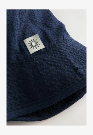 Tumesinine ämbri müts, mis on valmistatud tekstuuriga kangast, millel on õmmeldud muster ja ruudukujuline logo kinnitus koos päikesemustriga külgedel.