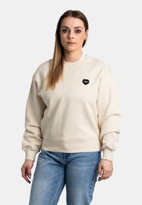 Lichtbeige sweatshirt met een ronde hals en omgeslagen mouwen, met een klein zwart hartvormig geborduurd logo op de borst.