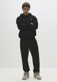 Black hoodie with a front pocket, matching black joggers, and grey sneakers. Model stands with arms crossed in a neutral setting.