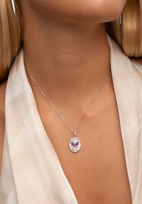 Collar de cadena de plata con un colgante ovalado que presenta un diseño de mariposa en tonos morado y rosa, exhibido sobre una blusa de color claro.