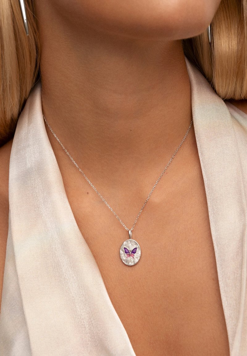 Collar de cadena de plata con un colgante ovalado que presenta un diseño de mariposa en tonos morado y rosa, exhibido sobre una blusa de color claro.