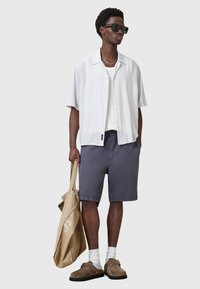 Camicia a maniche corte a righe bianche sovrapposta a una canotta bianca, abbinata a pantaloni corti blu-grigi. Scarpe slip-on marroni e borsa a tracolla beige. Occhiali da sole scuri.