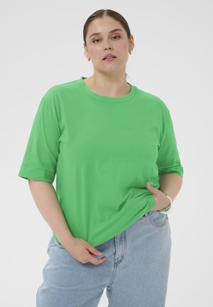 Femme aux cheveux attachés portant un t-shirt vert vif et un jean bleu clair, debout devant un fond blanc uni.