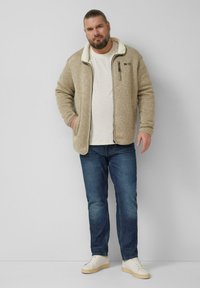 Mężczyzna w beżowej polarowej kurtce na białej koszuli, niebieskich dżinsach i białych trampkach stojący na tle jednolitego, jasnego tła.