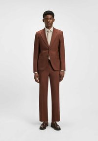 Brun skräddarsydd kostym med en kavaj med en knapp, matchande byxor och en krämfärgad skjorta med prickigt mönster. Mörka loafers fulländar looken.