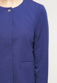 Fioletowa kurtka z płótna z okrągłym dekoltem, zapięciem na zatrzaski z przodu i pojedynczą naszywaną kieszenią po lewej stronie. Gładka tekstura.