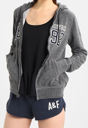 Personne portant un sweat à capuche gris zippé avec le numéro « 92 » et un short bleu marine avec l'inscription « A&F », associé à un débardeur noir en dessous.
