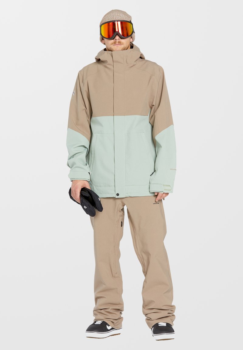 Lichte beige en mintgroene waterdichte jas met capuchon, gecombineerd met bijpassende beige broek en zwarte handschoenen, ontworpen voor wintersport.