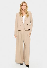 Completo beige su misura con giacca accorciata caratterizzata da doppi bottoni e pantaloni a gamba larga. Tessuto morbido con una vestibilità rilassata e pieghe sottili.