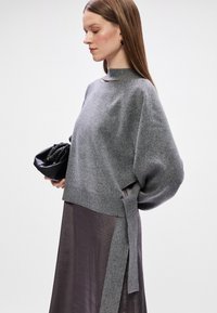 Γκρίζο πλεκτό πουλόβερ με υψηλή λαιμόκοψη, χαλαρή εφαρμογή και λεπτομέρεια με κορδόνι στο πλάι. Φορεμένο πάνω από μια σκούρα, φαρδιά φούστα και συνδυασμένο με μια μικρή μαύρη τσάντα.