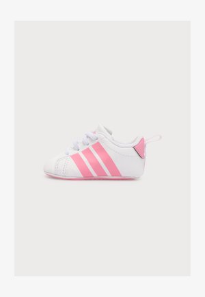 Biały dziecięcy sneaker z różowymi paskami, różowym zapiętkiem i białymi sznurówkami, pokazany z boku na jasnym, jednolitym tle.