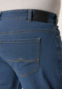Jeans de mezclilla azul oscuro con una etiqueta de cuero negro en la cintura, costuras en contraste y un bolsillo trasero con un detalle de puntada en zigzag.