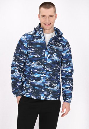 Blauw-patronen waterdichte jas met golvenontwerpen, een capuchon en elastische manchetten, gedragen met een wit shirt en zwarte broek.