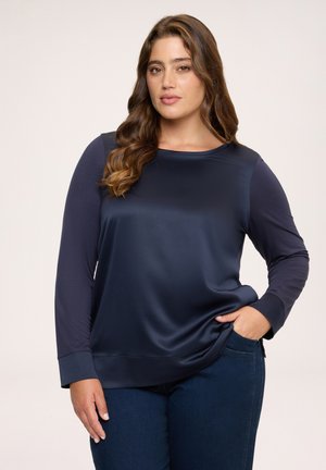 Kvinde med langt, bølget hår iført en mørkeblå satin top med lange ærmer og mørke jeans, stående med den ene hånd i lommen mod en ensfarvet baggrund.
