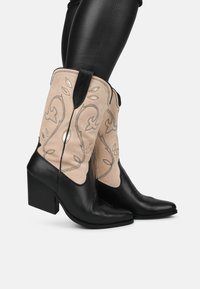 Botas de vaquero de cuero negro con una parte superior de ante color canela, que presentan un bordado intrincado, punta afilada y un tacón cuadrado. Detalles de patrón decorativo.