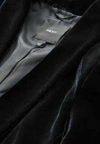 Blazer de terciopelo negro con una textura suave, que cuenta con un forro de satén gris y una etiqueta que dice "NEXT". Enfocado en el diseño del cuello y las solapas.