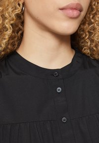 Μαύρη πουκάμισο με κολάρο και πτυχές, διαθέτοντας τρία μαύρα κουμπιά και λεία υφή. Εστίαση στην περιοχή του λαιμού και τις λεπτομέρειες της πουκαμίσας.