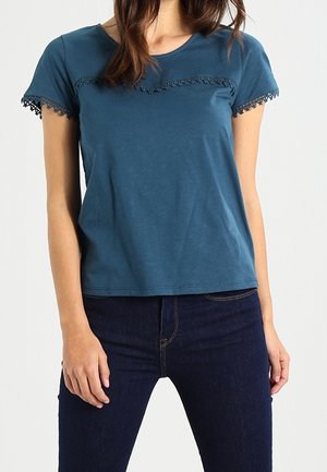 Vrouw draagt een blauw topje met korte mouwen en kanten rand op mouwen en borst, gecombineerd met donkerblauwe spijkerbroek tegen een effen achtergrond.