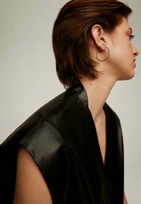 Prenda de cuero negro con un escote ancho en forma de V y mangas cortas. La modelo lleva un aro en la oreja y tiene el cabello corto y marrón.
