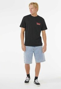 Jonge man in een zwart graphic t-shirt, lichtblauwe denim shorts, zwarte sokken en zwarte sneakers, staand en naar de zijkant kijkend.