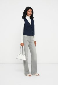 Kobieta w granatowej kamizelce, białym golfie, szarych dżinsach, beżowych sandałach na obcasie, trzymająca białą torebkę, stojąca na tle jednolitego białego tła.