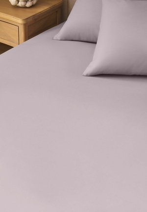 Letto con lenzuolo lavanda liscio e due cuscini abbinati accanto a un comodino in legno con oggetto decorativo.