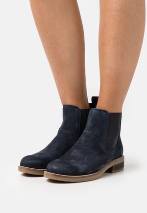 Botas de tobillo de ante azul oscuro con paneles laterales elásticos y suelas marrón claro, utilizadas sobre piernas desnudas contra un fondo blanco.