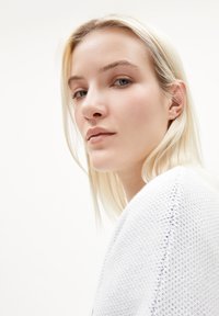 Nahaufnahme einer Person mit hellblondem Haar, die ein strukturiertes weißes Kleidungsstück trägt. Der Hintergrund ist schlicht und neutral, wodurch die Gesichtszüge betont werden.