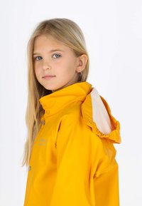 Jeune fille aux longs cheveux blonds portant un imperméable jaune vif avec une capuche sur un fond blanc uni.