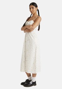 Vestido midi floral con tirantes finos, tejido color crema claro, cinturilla elástica y pequeños patrones de flores en tonos pastel. Zapatos negros y calcetines blancos.