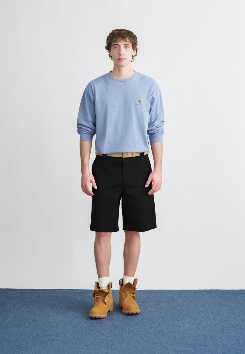 Camicia blu a maniche lunghe, pantaloncini neri, stivali da lavoro color cuoio, con calzini bianchi. Presenta un logo sulla camicia e una cintura con una fibbia semplice.