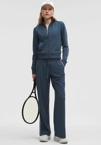 Plavi sportski trenerka s bijelim prugama, koja uključuje duksericu sa patentnim zatvaračem i hlače s vezicom, uparen s teniskim reketom.