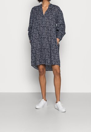 Nainen, jolla on yllään tummansininen pitkähihainen mekko, jossa on valkoinen paisley-kuvio ja valkoiset tennarit, seisoo yksinkertaista taustaa vasten.